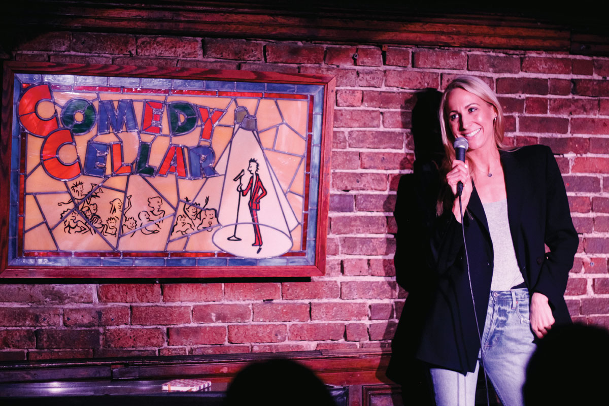 Nikki Glaser performing stand-up at Comedy Cellar. She is a blond woman wearing a black blazer and jeans. She is holding a microphone and standing close to a wall of bricks and a sign of mosaic glass that reads "comedy cellar."