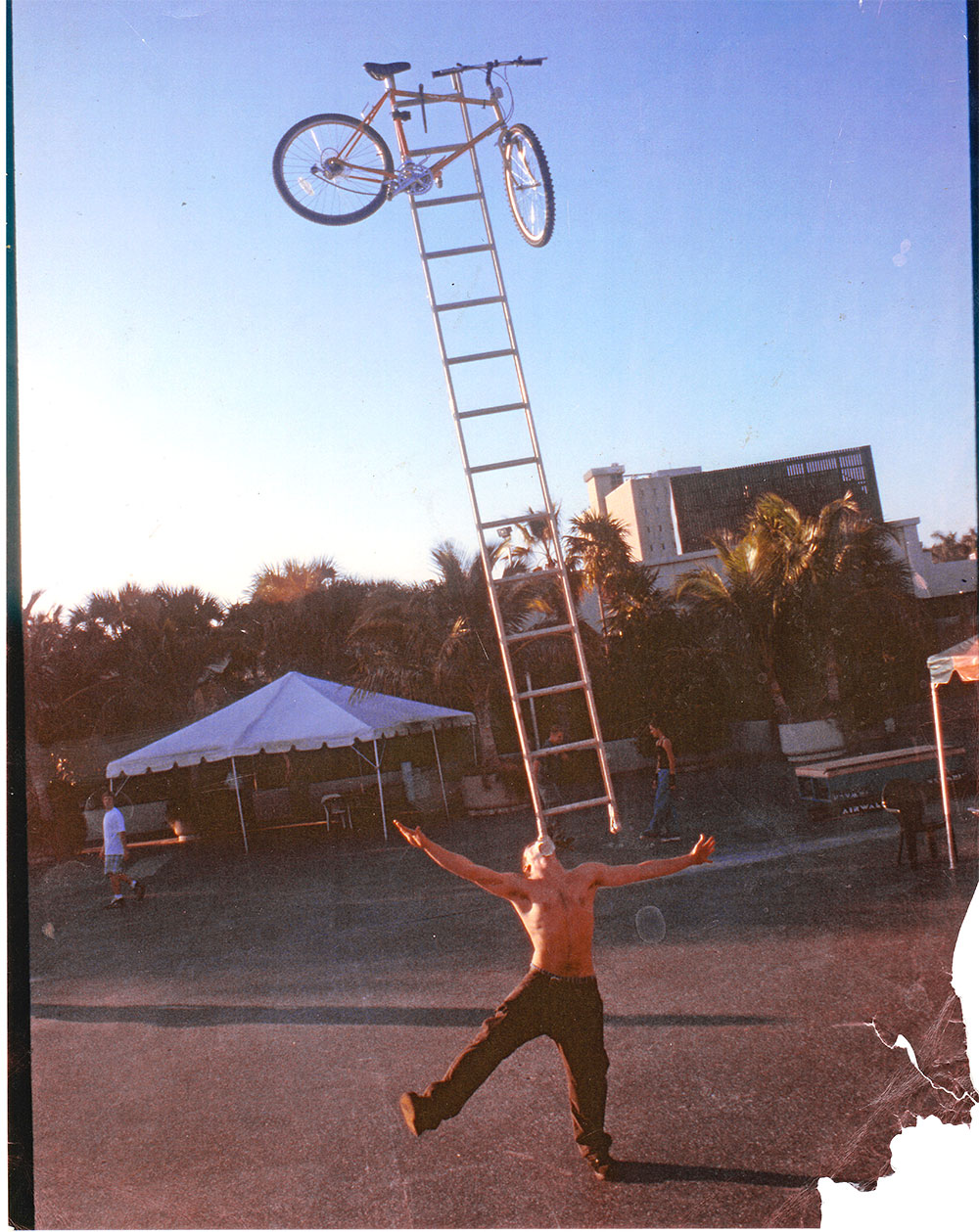 A picture of Steve-O balancing ladder and bike on his chin. A shirtless white man with one foot on the ground and arms open wide balances a bike on top of a ladder on his chin.
