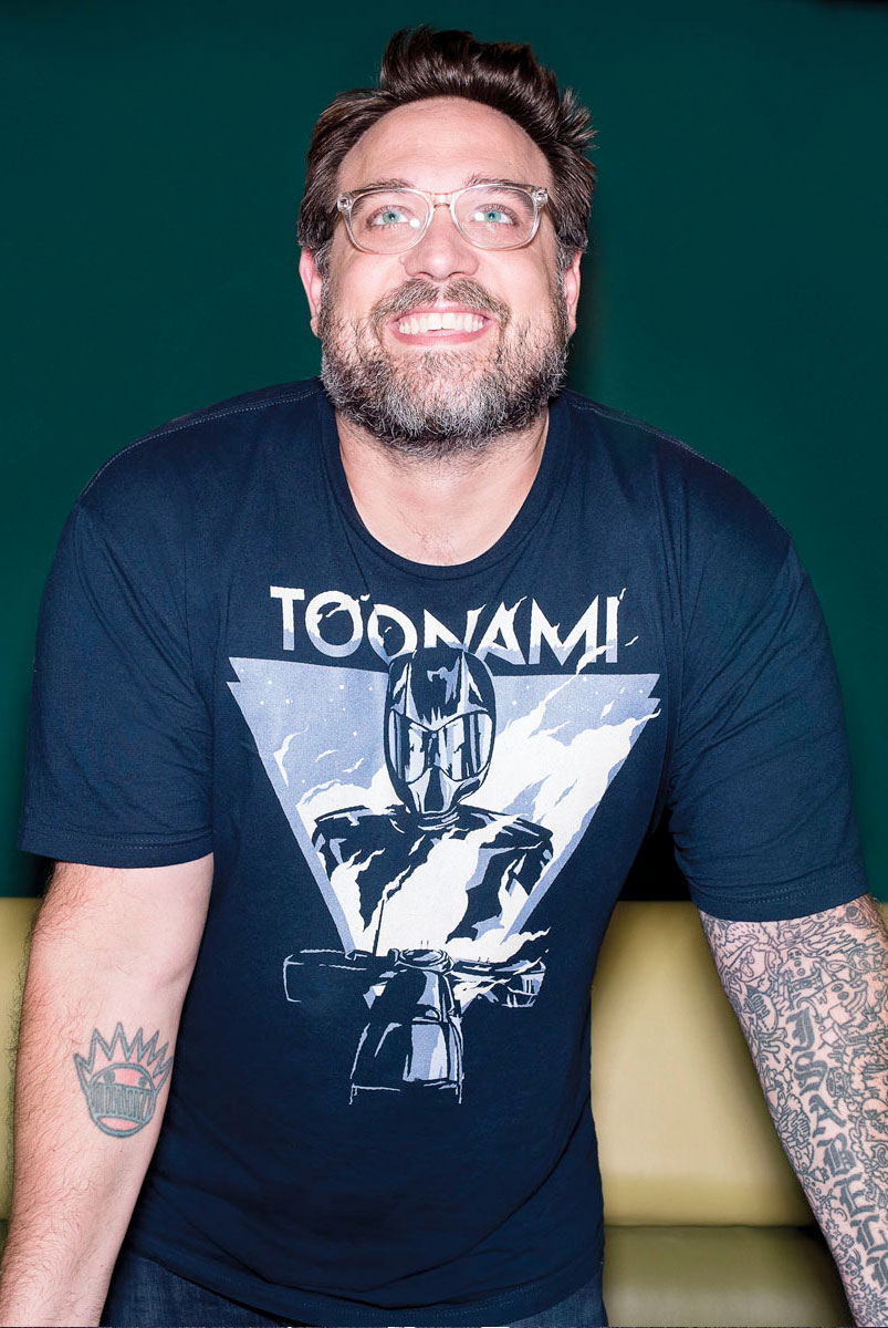 Jason DeMarco stands, leaning toward the camera, looking upward. He wears glasses, a scruffy beard, and a toonami shrit. 