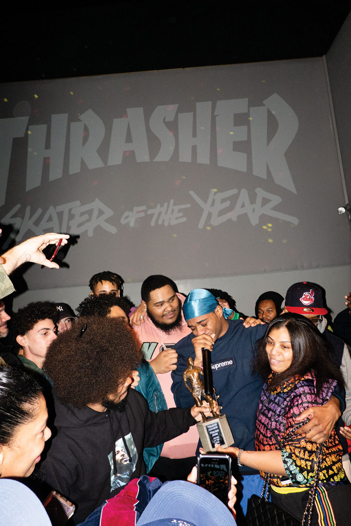 Atiba Jefferson's photo of Thrasher, the skater of the year. A group of people hug and stand around a man with a do rag and a microphone. In the background, projected on the wall words read thrasher, skater of the year.