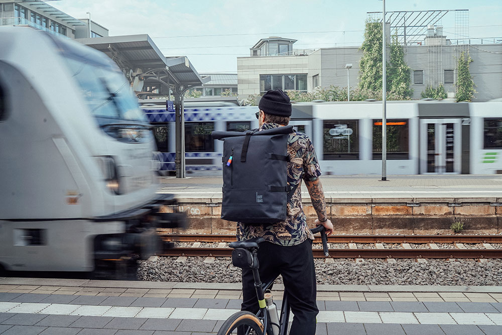 Person riding bike in city wearing GOT BAG Rolltop bag. The man wears a beanie, sunglasses, and a floral shirt. His arms are tattooed and he's waiting on a subway platform as a train passes by.