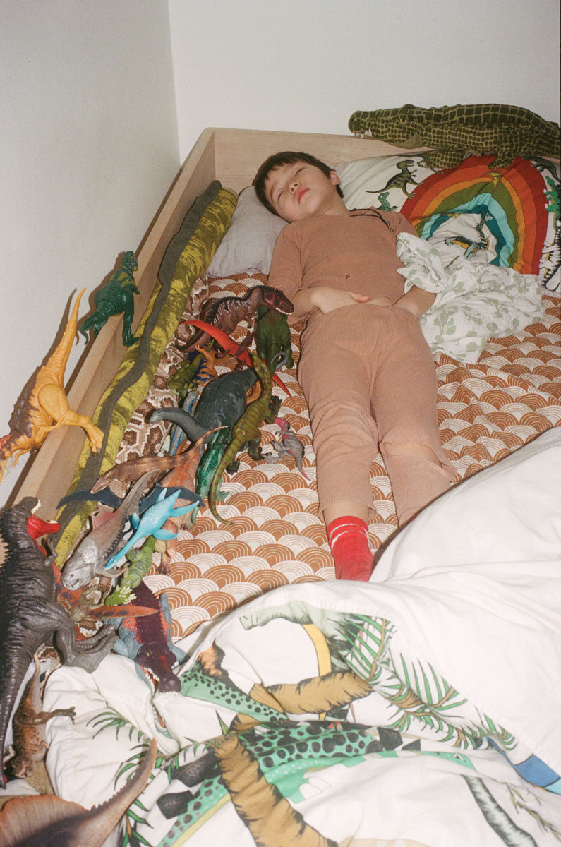 A small child in beige pajamas sleeps in a kid's bed surrounded by pillows, blankets and many many plastic dinosaur toys of all shapes, types, and sizes. 