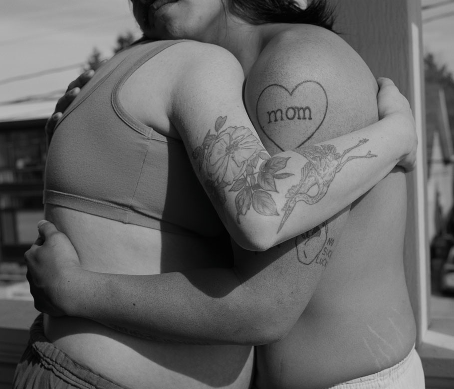 A black and white photograph of two people hugging. One wears a sports bra, the other no shirt. The arms nearest each feature a tattoo.