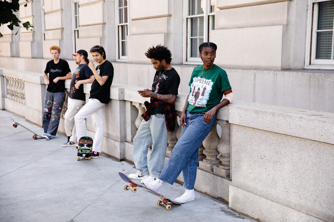 Atiba Jefferson's photo of skate session group. Five people wearing jeans and shirts lean against a concrete wall. Three of them rest their foot on skateboards on the ground. 