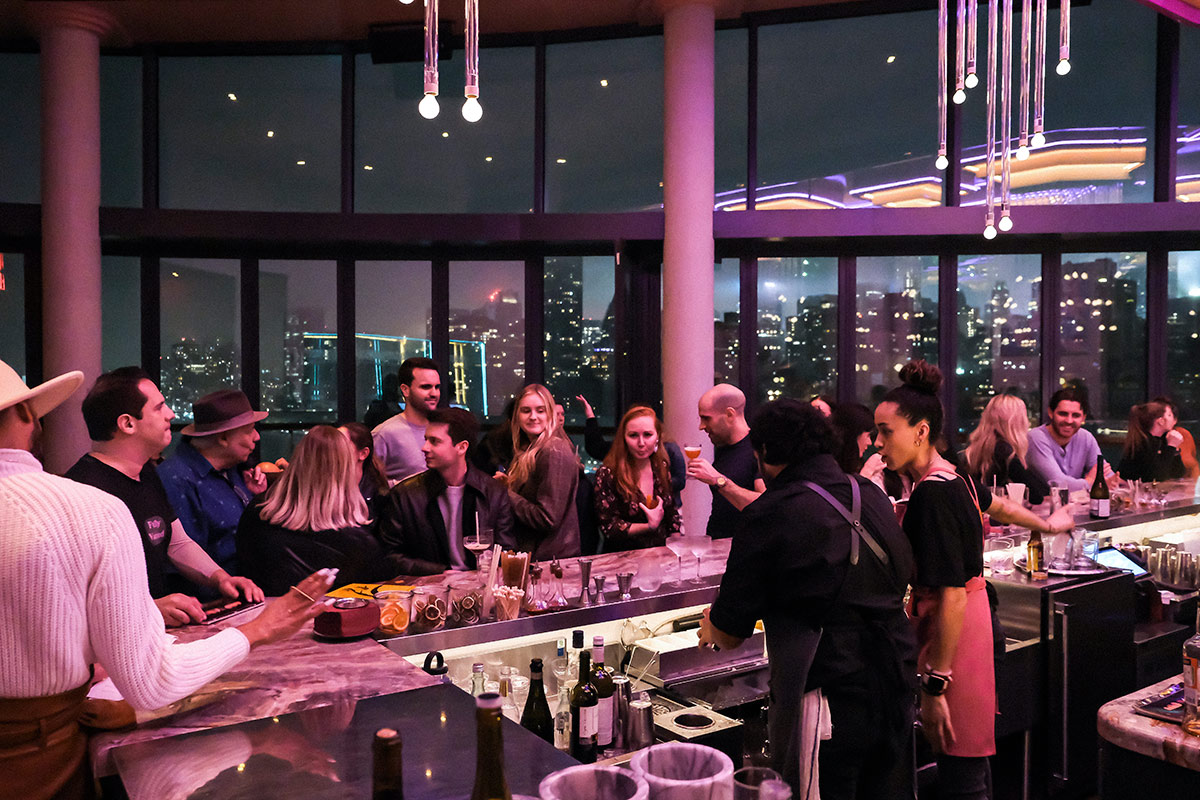 Many people gather at the bar of the Panorama Room, the rooftop bar at The Graduate Roosevelt. They are lit by pink and purple light and behind them is a wall of windows overlooking new york city.