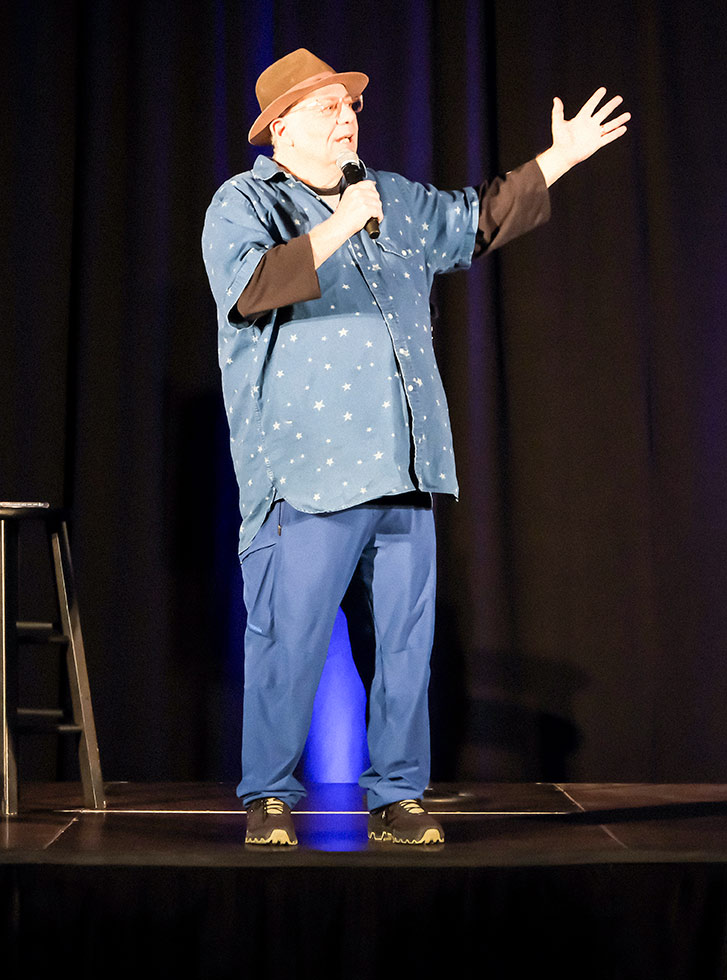 Eddie Pepitone performs on a stage at the Extra Credit Event Series by Graduate Hotels and Whalebone Magazine. He's holding a microphone and wears an orange fedora and stands in front of a stool.