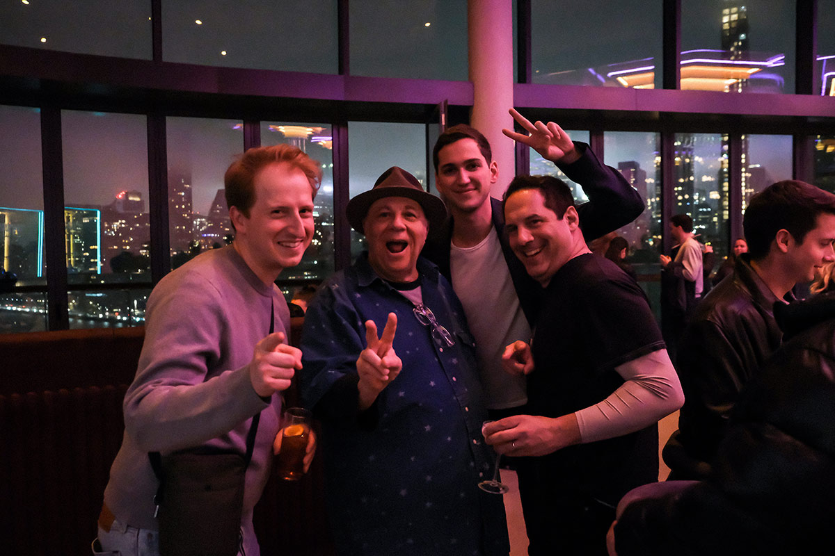 James Austen Johnson, Matt Friend, Seth Herzog and Eddie Pepitone lean in together for a group photo and smile at the camera. In the background is a wall of windows over looking a city at night.