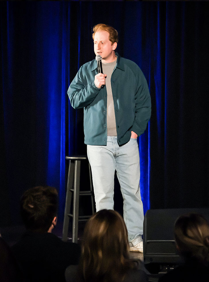 James Austin Johnson performs on a stage at the Extra Credit Event Series by Graduate Hotels and Whalebone Magazine. He's holding a microphone and wears a green jacket and light pants and stands in front of a stool.