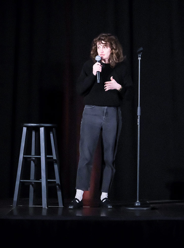 Jo Firestone performs on a stage at the Extra Credit Event Series by Graduate Hotels and Whalebone Magazine. She's holding a microphone and wears a long sleeve black shirt and dark pants and stands in front of a stool.