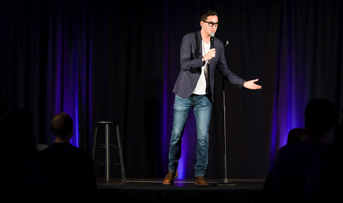 Matt Friend performs on a stage at the Extra Credit Event Series by Graduate Hotels and Whalebone Magazine. He's holding a microphone and wears a dark blazer, white shirt, and jeans and stands in front of a stool.