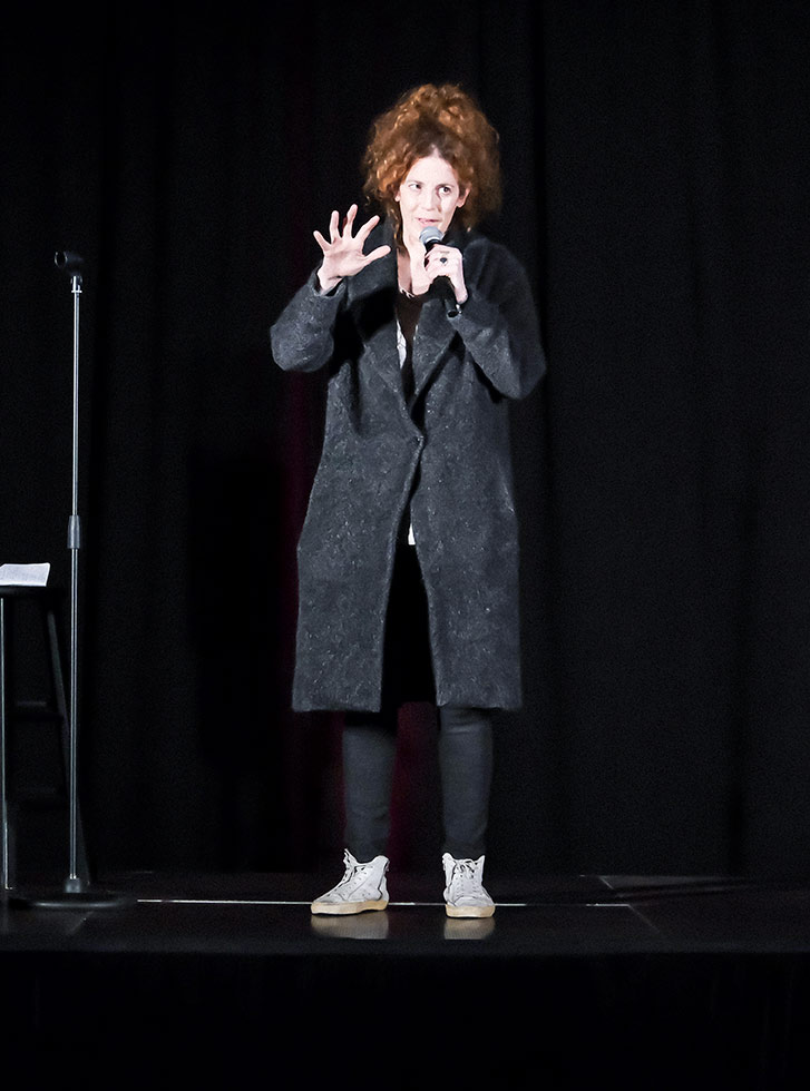 Morgan Murphy performs on a stage at the Extra Credit Event Series by Graduate Hotels and Whalebone Magazine. She's holding a microphone and wears a long dark coat and stands in front of a stool.