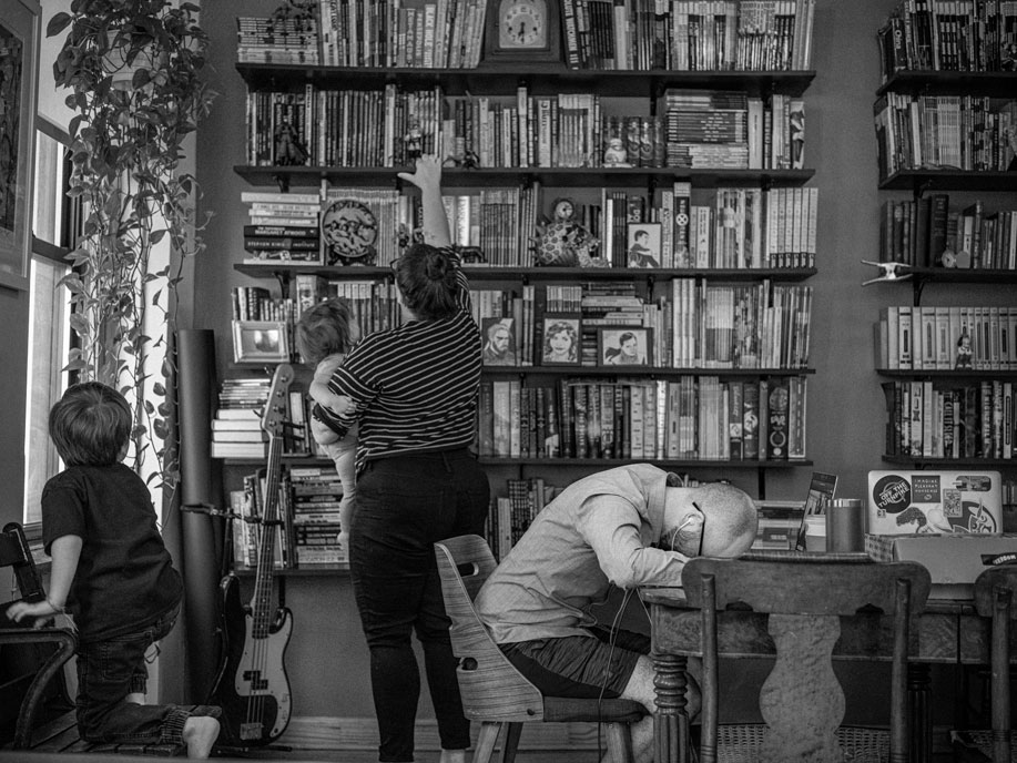 A woman holding a child reaches for a high shelf amongst a wall of books. In the fore ground a man sits with his head on top of a table. A small child, his face turned away, watches the woman. 