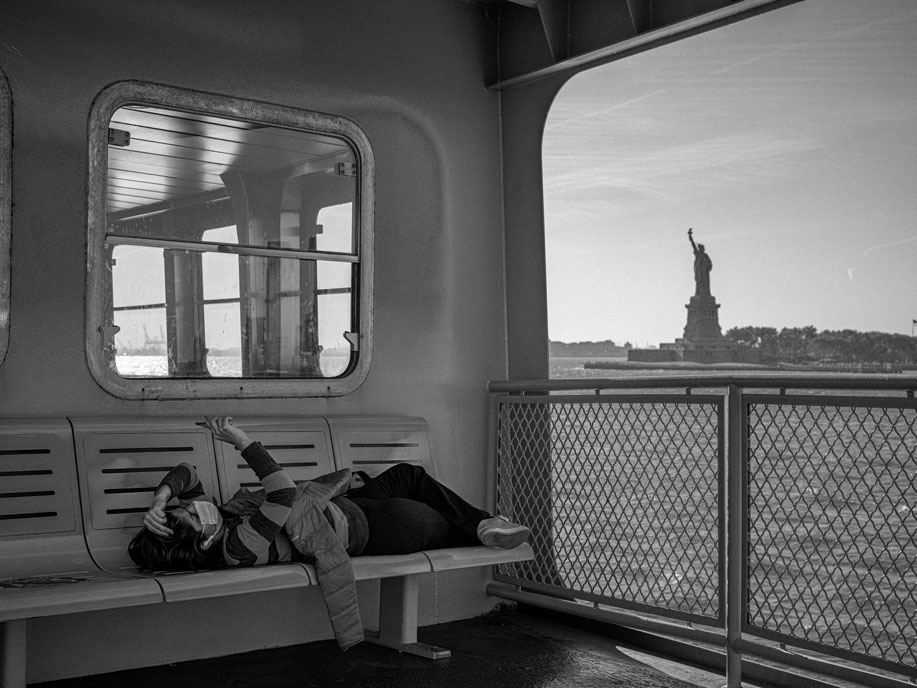Black and white photography of a masked person laying across a row of seats on a ferry. In the background is water and the statue of liberty.