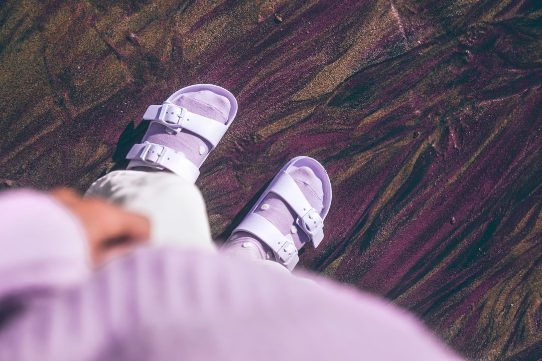 Photograph of someone wearing purple sandals standing on purple sand.