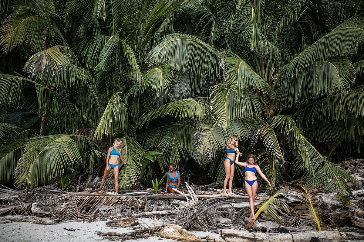 Four women stand amongst plant and debris under long green palms. The women wear blue bikinis and are facing outward tot the shore.