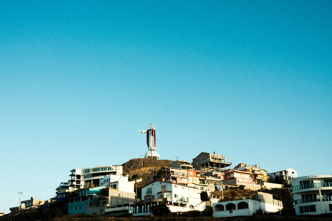 Blue sky fills the photo. In the lower half, a large state of jesus stretches their arms out and over a hill covered in houses. 