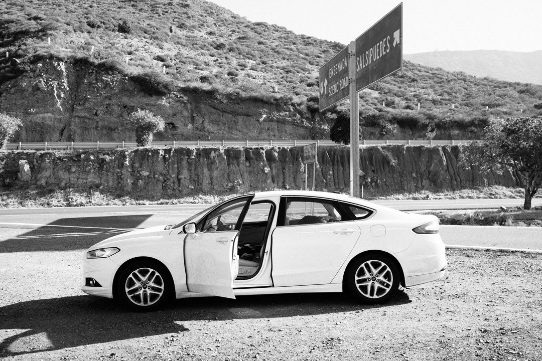 A white sedan sits against a hilly desert back drop. The driver doors hangs open and the car is empty.