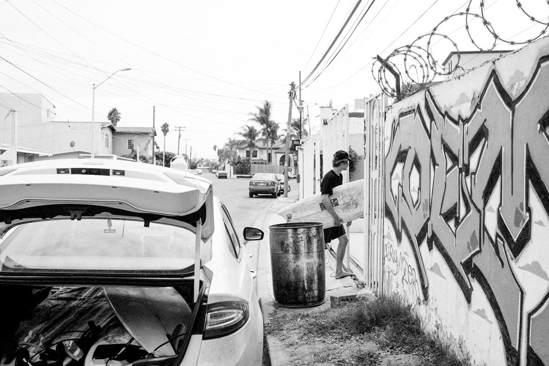 A white sedan is parked next to a concrete wall decorated with graffiti and adorned by barbed wire. A man dress in black with a baseball cap holds a surfboard and is entering into an opening of the wall.