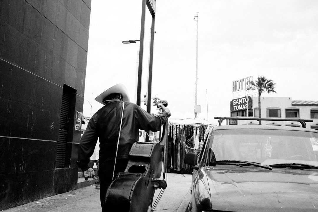 A man holding a large double bass and wearing a cowboy hat walks forward past a parked car.