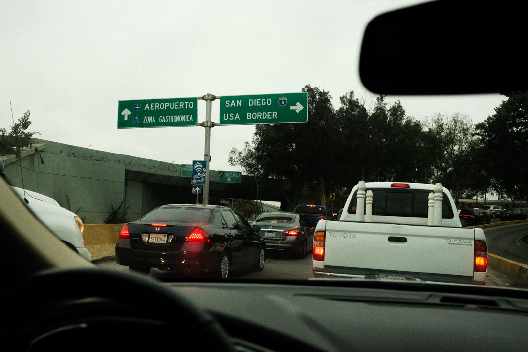 Looking through the windshield of a car. Two lanes of traffic stretch ahead. Signs pointing to the the US Mexico border.