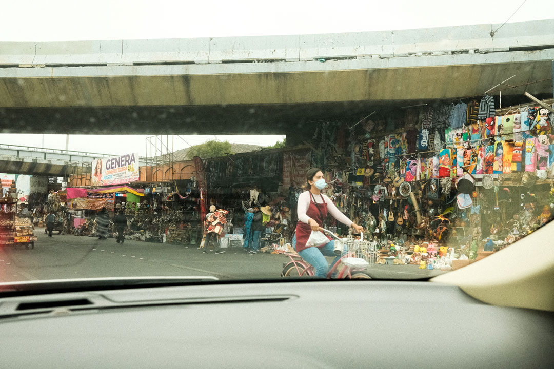 Looking through the windshield of a car. In the background a woman wearing a mask and an apron rides her bike. Behind her are stands of arts and touristy goods underneathe the bridge.