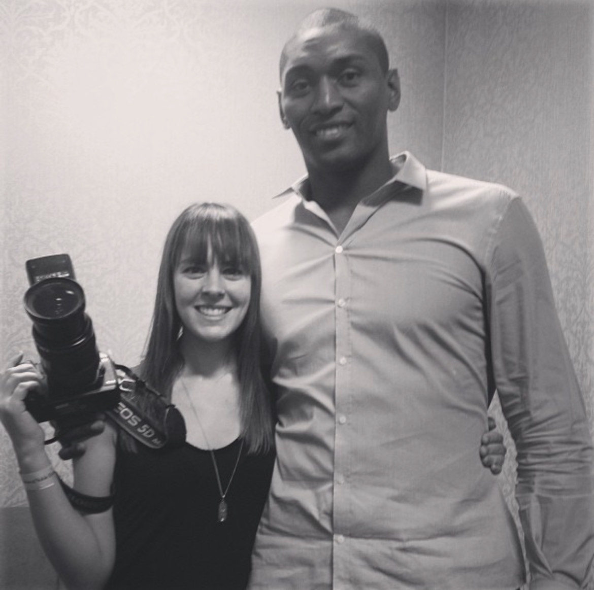 Image of Laura June Kirsch and NBA player Ben Simmons. Laura is a shorter white woman, wearing a black tank top and holding a large canon camera with flash. Ben is a tall black man wearing a light collared shirt. They're both looking forward and smiling.