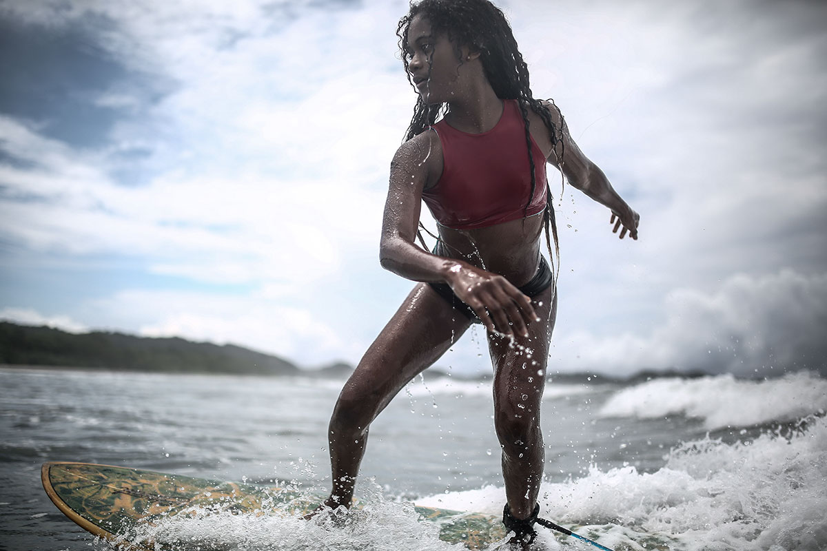 A young girl leans forward while standing on her surf board in a small swell.