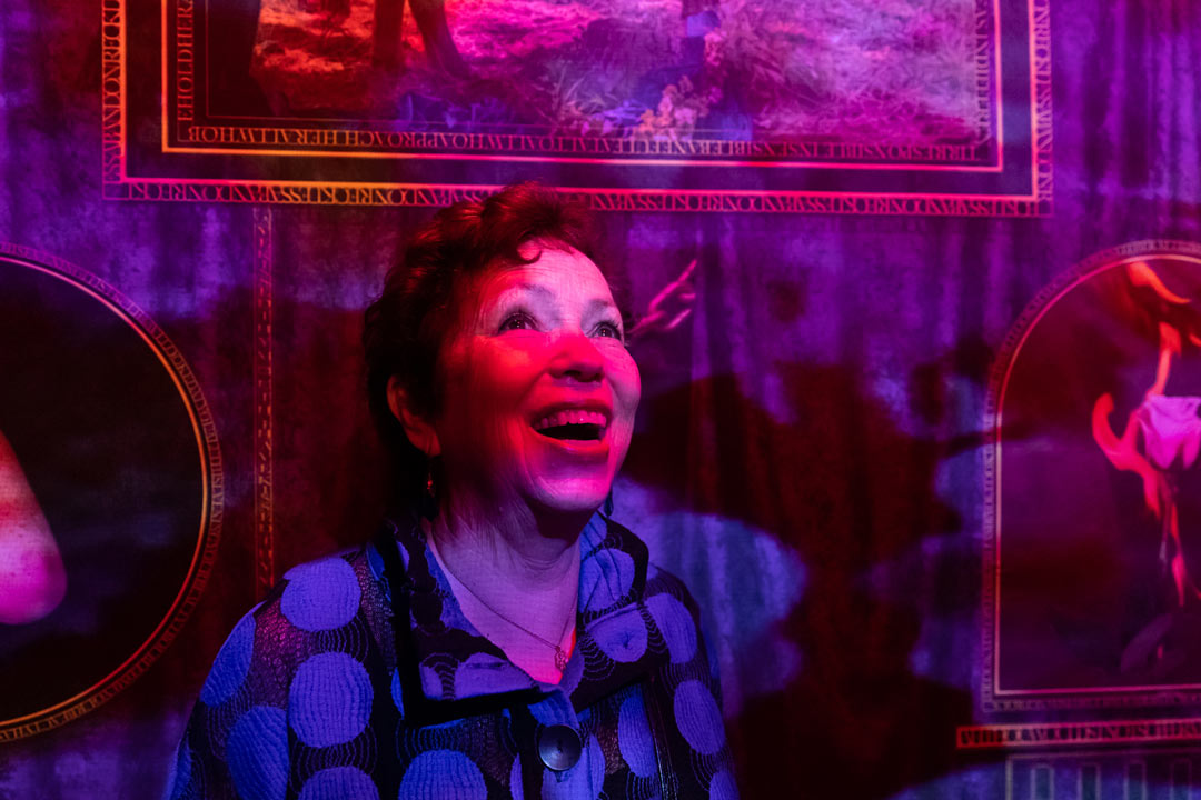 Close up photo of Meryl Meisler on her way into the club, covered in pink and purple lights, as she stops and smiles while looking towards the top of the stairs.