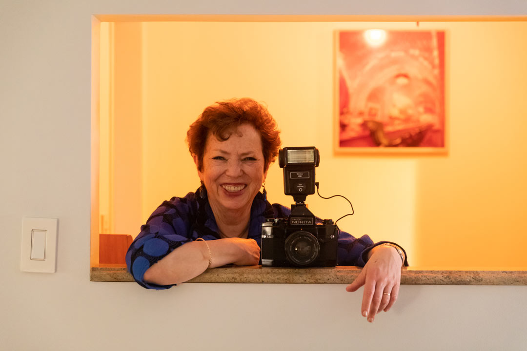 Photo of Meryl Meisler in her apartment smiling for a picture while posing with her camera.