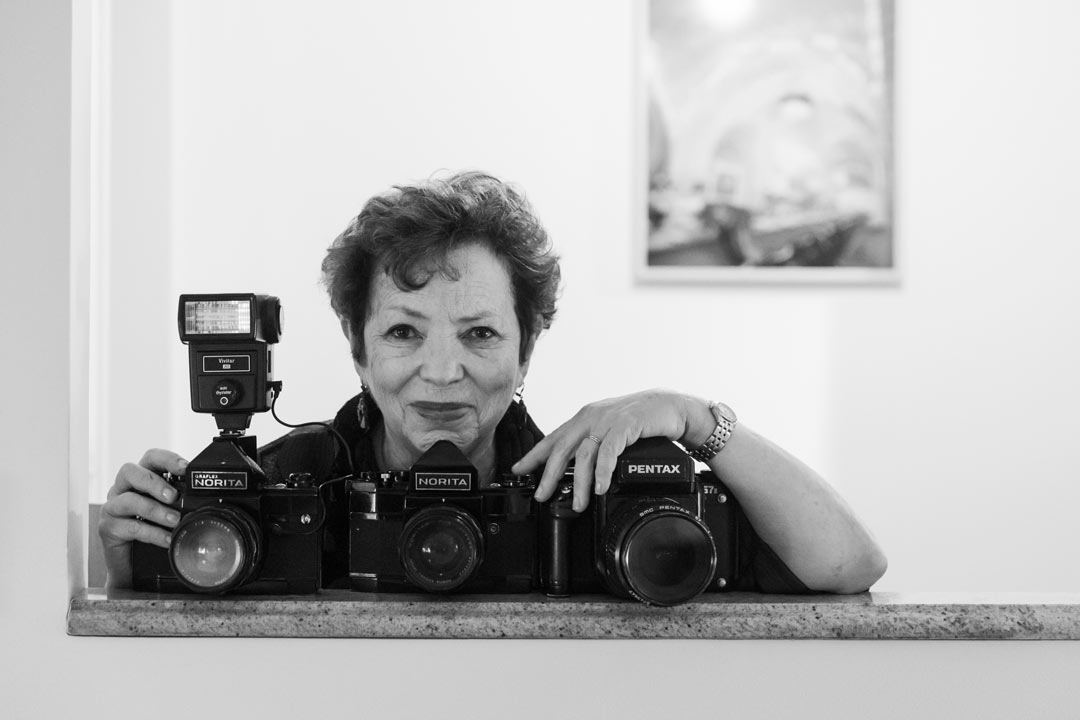 Black and white photo of Meryl Meisler in her apartment posing for a picture with three of her cameras.