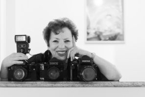 Black and white photo of Meryl Meisler in her apartment smiling for a picture with three of her cameras.