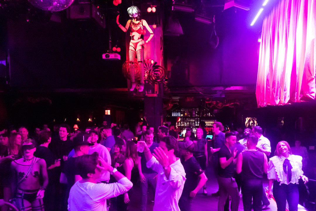 Photo of a large group of people mingling in the middle of the club covered in pink and purple lights. Hanging above them in the middle is a large mannequin body wearing a bikini and mesh tights.