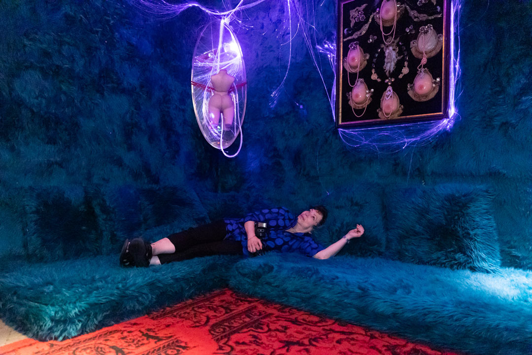 Photo of Meryl Meisler in the fuzzy room laying on a turquoise-green fuzzy couch. On the wall above her is a naked mannequin body and a picture frame holding six silicone boobs in a shrine-like fashion.