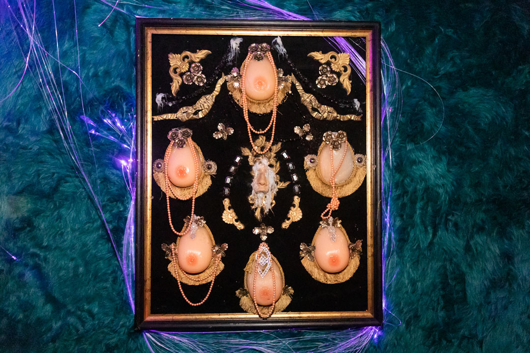 Close up image of the picture frame with six silicone boobs arranged in a circle adorned with beads and jewels. In the middle is a mold of the nose and upper lip from someones face.