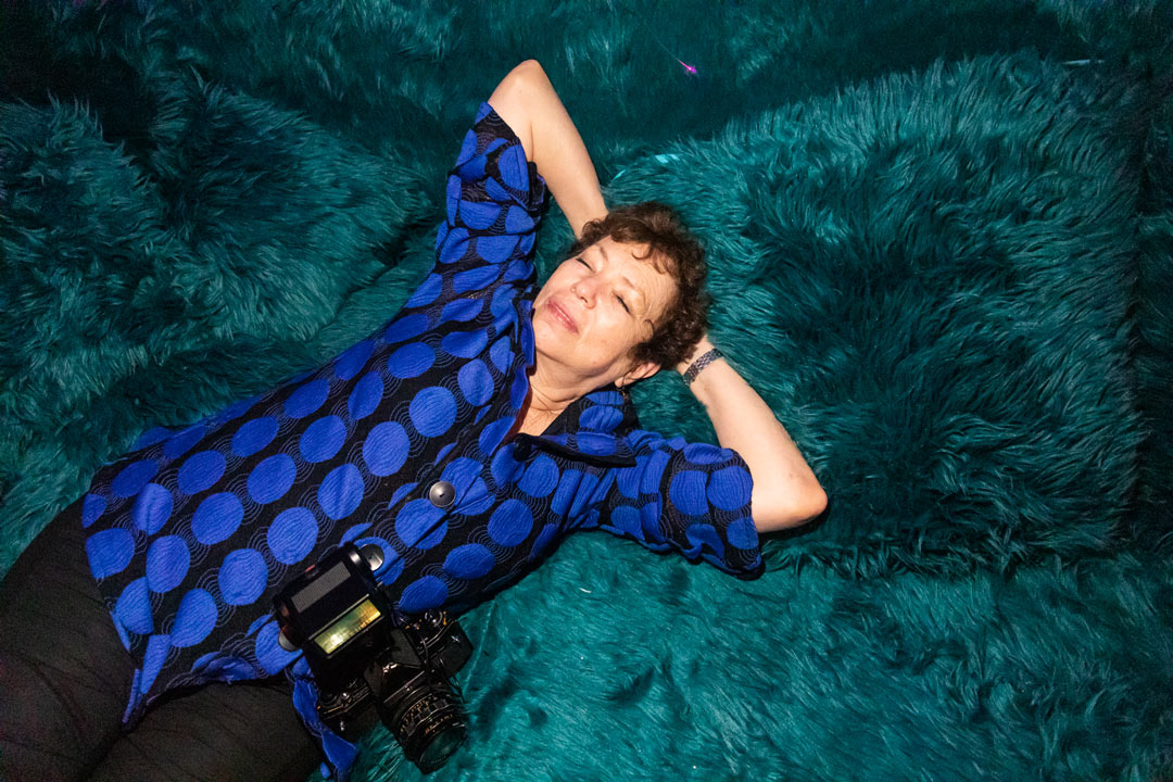 Photo of Meryl Meisler laying on a turquoise-green fuzzy couch with her hands behind her head and camera draped around her neck.