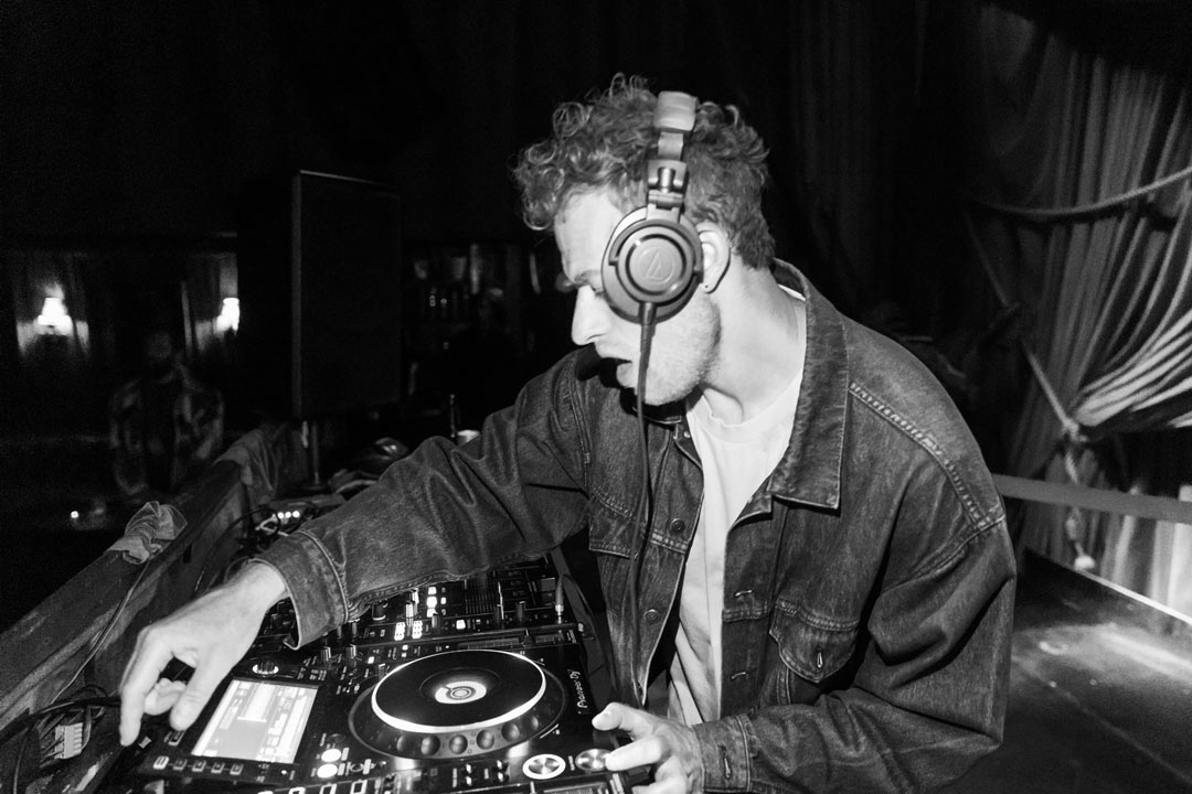 Black and white photo of DJ Child Of wearing a demin jacket and large headphones over his ears. He's holding the mix table and about to press one of the buttons of the machine.