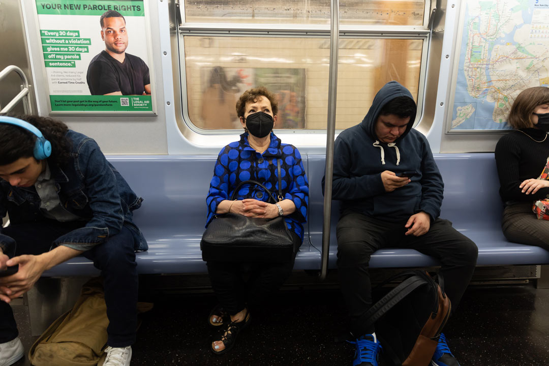 Photo of Meryl Meisler riding the subway, sitting on a blue bench and wearing a mask in between two strangers.