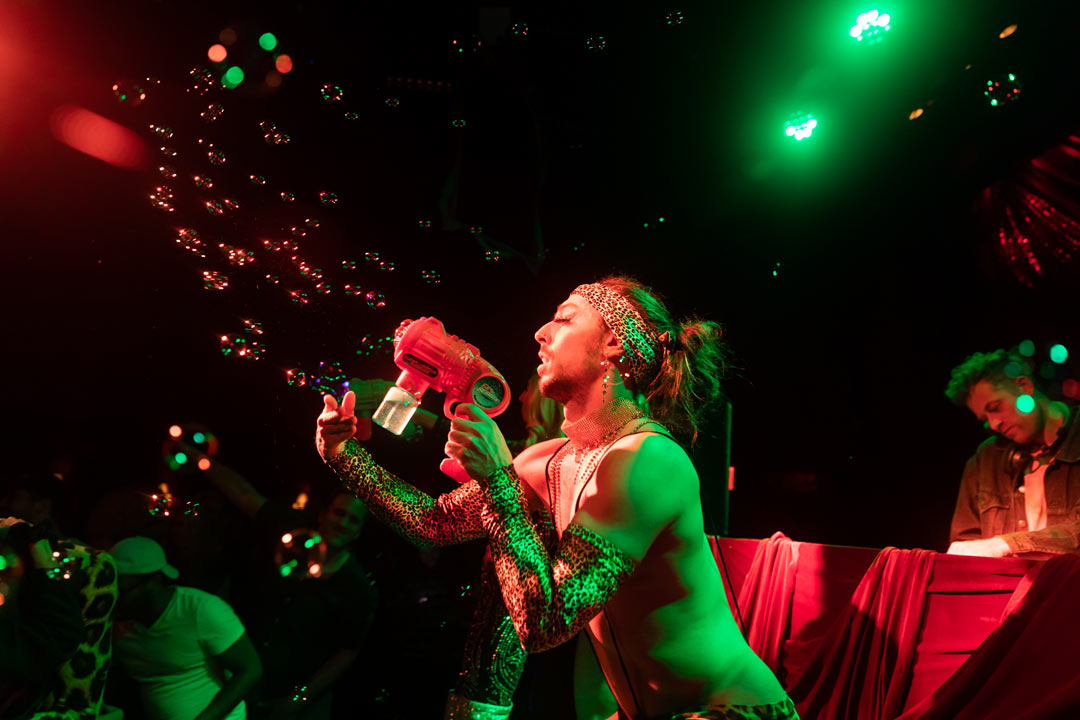 Photo of a topless club performer blowing bubbles out of a red bubble gun towards the crowd.