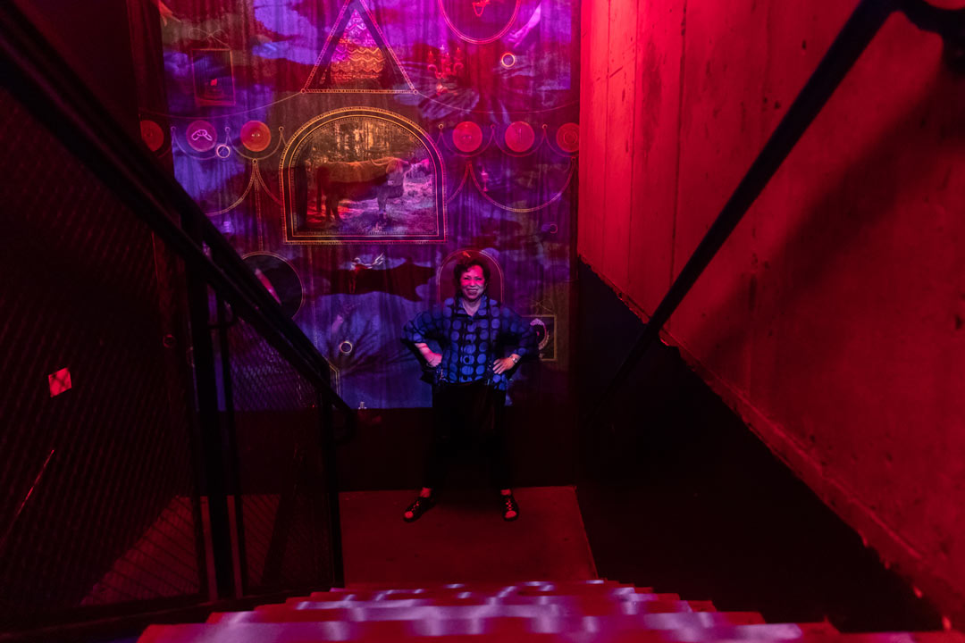 Photo of Meryl Meisler heading down into the nightclub covered in pink and red lights. She stops halfway down the stairs to turn and strike a pose wither her hands on her hips.