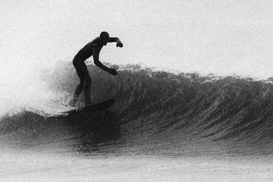 Black and white photo of a surfer in a wetsuit riding on a wave as it arches.