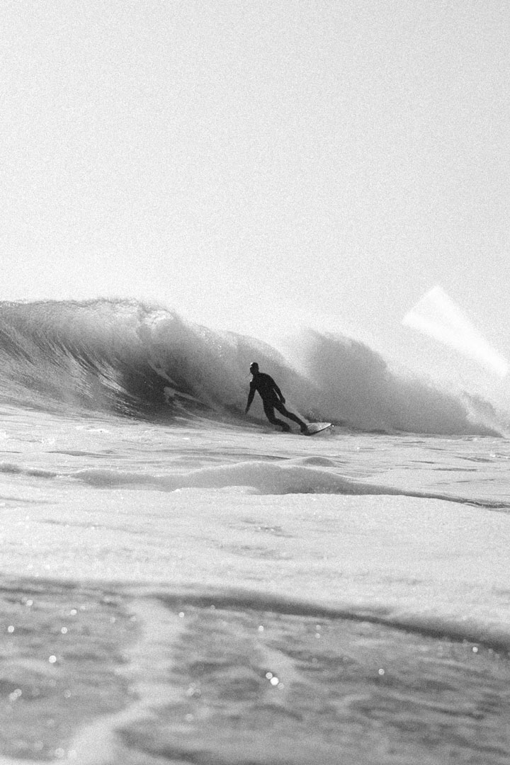 Black and white image of a surfer riding a wave as it crashes down behind them.