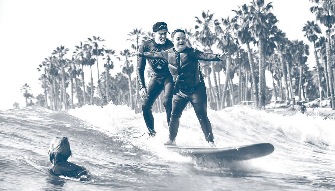 Photo from the Walk on Water foundation of an instructor tandem surfing with a special needs child while they cruise by someone swimming in the water.