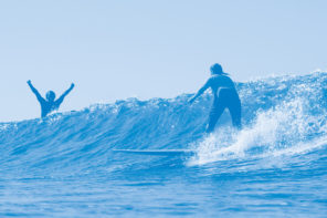 Photo of two surfers, one is riding the wave while the other is behind the crashing wave cheering with their arms in the air. The image has a blue colored overlay.