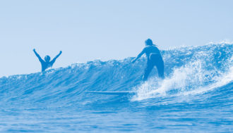 Photo of two surfers, one is riding the wave while the other is behind the crashing wave cheering with their arms in the air. The image has a blue colored overlay.