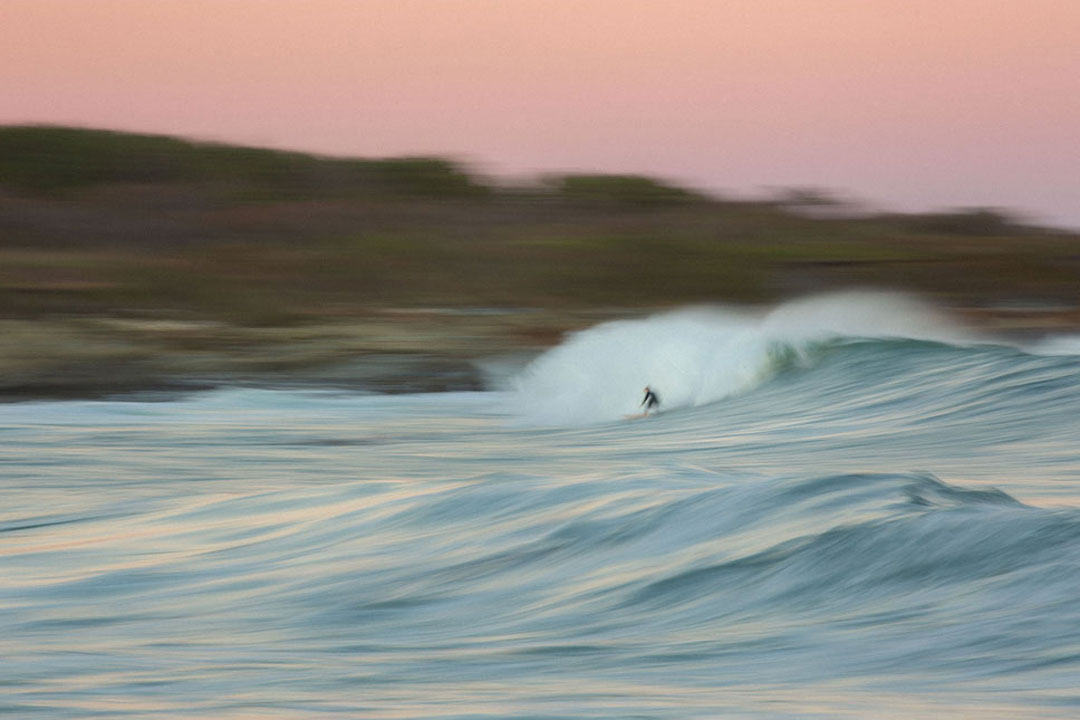 Blurred shot of a single surfer riding a large wave that looks like a black spec against the ocean. The pink sky contrasts against the blue ocean.