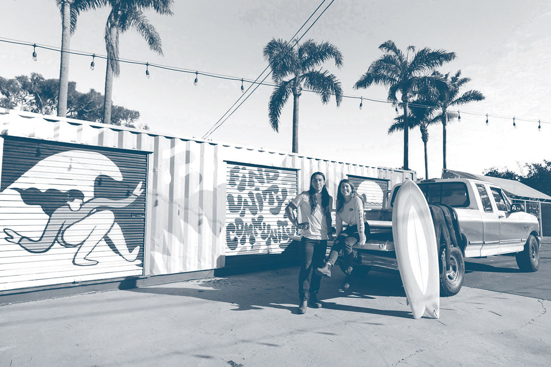 Photo of the co-founders of Un Mar de Colores, Mario Ordonez-Calderon and Kat Reynolds, sitting in the bed of a truck with a surfboard next to them. They're in front of garage doors with surf murals painted on them.