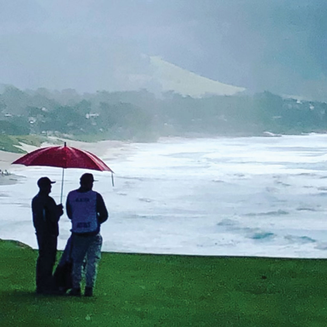 Kelly Slater and Benji Weatherley at a golf course by the ocean, standing under an umbrella.