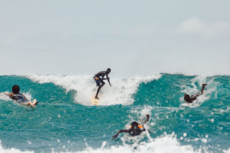 Photo of a group of surfers in the ocean paddling into a bright blue wave as the surfer in the middle jumps up to ride it.