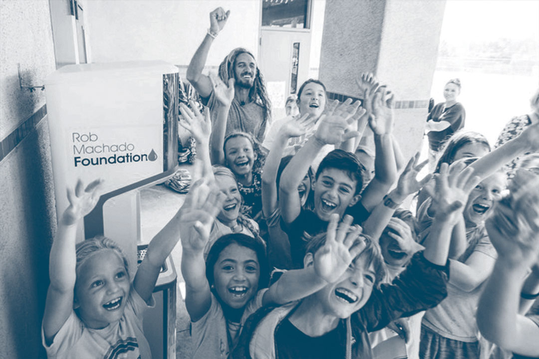 Photo from the Rob Machado foundation of a group of young kids smile and wave their hands in the air towards the camera with Rob Machado in the background.