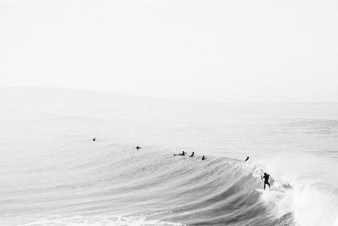 Black and white photo of a group of surfers paddling over a wave while one rides the wave on the far right of the image.